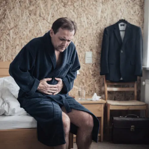 Blasser Mann im Bademantel sitzt vornübergebeugt auf der Bettkante und hält sich krampfartig den schmerzenden Bauch.